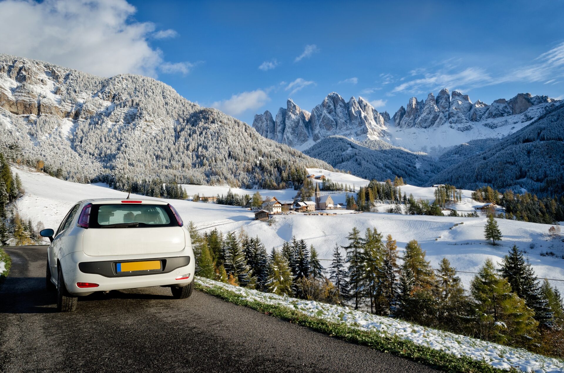 Voiture blanche sur la route lors d'un road trip dans un paysage des Dolomites en hiver avec de la neige sur les arbres, les montagnes de Geisler et l'église du village de St. Magdalena ou Santa Maddalena dans le Villnoesstal (Val Di Funes Valley) dans le sud du Tyrol, en Italie.