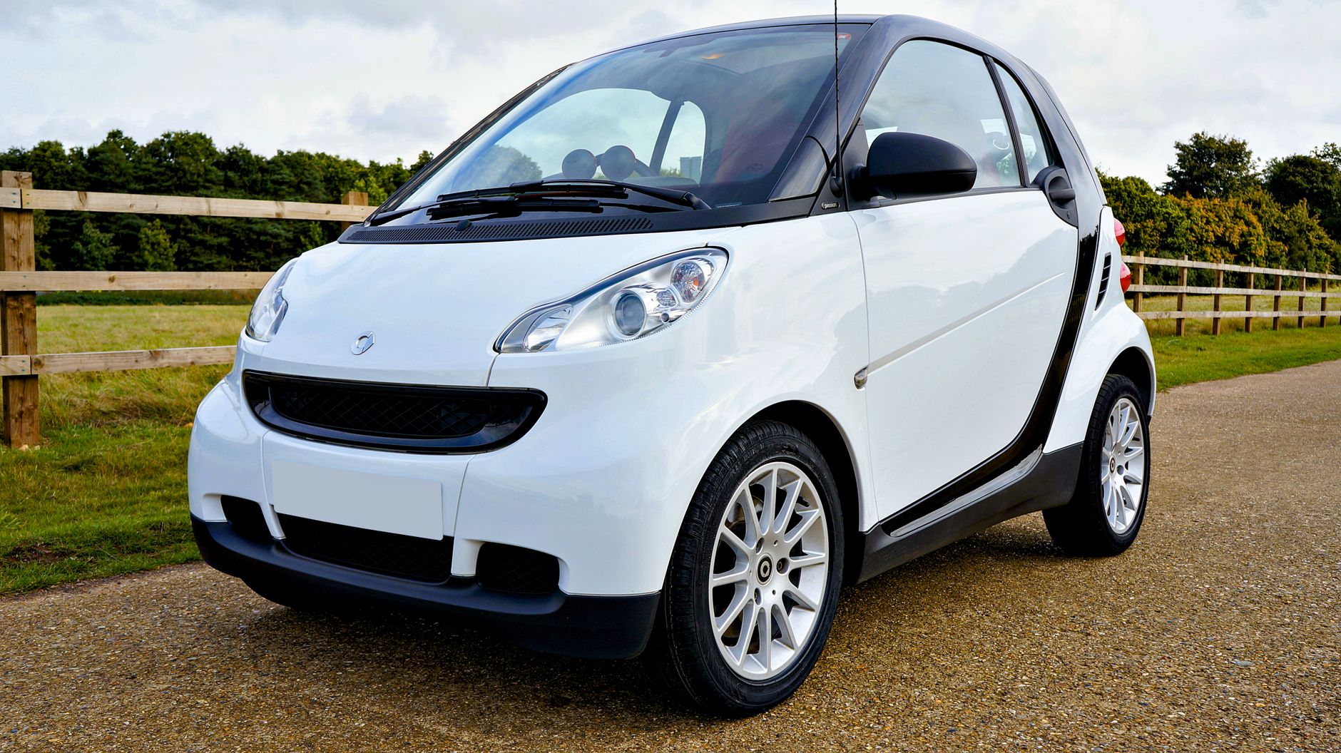 Une voiture Smart Fortwo blanche garée à l'extérieur par une journée ensoleillée avec verdure et clôture en bois.