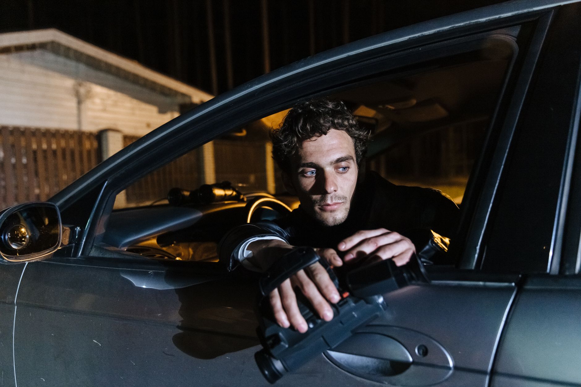 Un homme assis dans une voiture la nuit, tenant une caméra vidéo et regardant dehors.